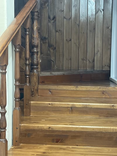 The image shows a wooden staircase with a dark wooden paneled wall behind it. The staircase has several steps, each made of polished wood. The banister is also made of wood, with decorative turned spindles. The background wall consists of vertical wooden planks, some of which appear to be stained darker than others. There is a white wall or object visible to the far right. The scene depicts an indoor setting, likely a residential interior. The specific location in Nachalovo, Russia, cannot be determined from the visual information provided in the image.