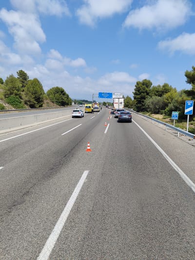 This is a daytime, eye-level, medium shot of a multi-lane highway in Altafulla, Spain, under a partly cloudy blue sky. The highway has multiple lanes with white lane markings. Several vehicles are present, including an ambulance, a white SUV, and a dark-colored car. There are orange traffic cones on the road, suggesting a traffic incident or road work. In the background, there are trees and vegetation on either side of the highway. Road signs indicating directions and distances are visible, including one for Barcelona and Lleida, and another for E-15 AP-7 KM 233, with a "100 m" sign. The overall scene depicts a typical highway environment with signs of temporary traffic management.