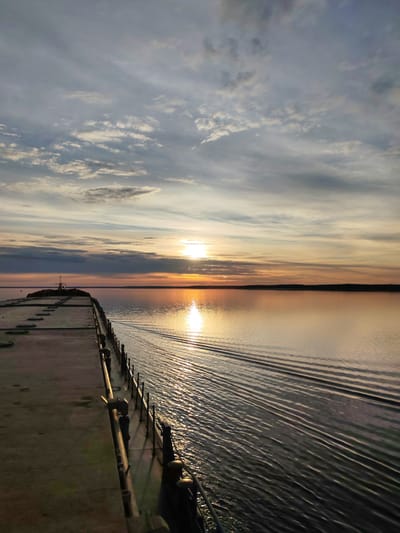 The image depicts a concrete pier extending into a calm body of water at sunset, with the sun's reflection shimmering on the water's surface. The sky is filled with scattered clouds, tinged with hues of orange, pink, and gray. A faint landmass with a dark silhouette is visible on the horizon. The pier itself is weathered, with visible seams and some metal railings along its edge. The scene is devoid of any people or animals, presenting a serene and isolated environment. The location is identified as Gorushka, Russia.