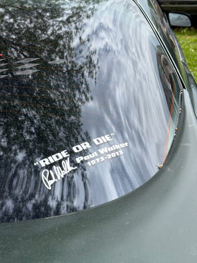 The image shows the rear window of a dark-colored vehicle. A white sticker with text is applied to the window. The text reads: "RIDE OR DIE" above a signature that appears to say "Paul Walker" and below that "1973-2013". The sticker is white and in a stylized font. Reflections of trees and the sky are visible on the car window, suggesting an outdoor setting. The bottom portion of the image shows the dark green textured surface of the car's exterior. The image was taken in Reckange-sur-Mess, Luxembourg.