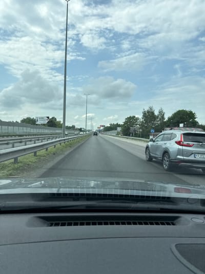 The image presents a daytime view from inside a vehicle, looking forward through the windshield onto a multi-lane highway under a partly cloudy sky. The sky is light blue with scattered white cumulus clouds.

In the mid-ground, a multi-lane asphalt highway extends into the distance. On the left side of the road, a metal guardrail runs parallel to the direction of travel, beyond which a concrete noise barrier wall is visible, topped by green trees. Several streetlights line the highway, including a prominent tall, grey pole in the left-center of the frame. Further down the road on the left, a white billboard with Cyrillic text, including "БУДИНКИ" and "СТРОИТЕЛЬ" (suggesting "buildings" and "builder/construction"), and a right-pointing arrow, is positioned above the noise barrier. Another multi-lane section of the highway with ongoing traffic is visible to the left.

On the right side of the road, a silver Honda CR-V (likely 5th generation) with a partially visible roof cargo carrier or tent, covered by a patterned tarp, is driving in the same direction as the viewer. The vehicle's license plate, "KA 4932 I," is clearly visible, identifying it as registered in the Kyiv region of Ukraine. Further ahead on the right, additional trees and a building with a striped roof are visible, along with a blue circular traffic sign depicting a white right-pointing arrow. Multiple vehicles are observed in the distance in both directions of travel.

The foreground of the image reveals the interior dashboard and windshield base of the viewer's vehicle, showing air vents and reflections on the glass.

Based on the visible Ukrainian license plate from the Kyiv region, and in consideration of the user's specific query, the scene is located in Ukraine, on a major highway such as Peremohy Street in Kyiv, given its capacity to be a multi-lane thoroughfare matching this description. No people are distinctly visible in the frame.