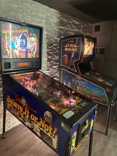 The image displays an indoor setting, likely a game room or private arcade in Winterthur, Switzerland, featuring three pinball machines. The room has textured, light-colored walls resembling stacked stone and a warm-toned wooden plank floor. The lighting is dim, creating an atmospheric ambiance typical of game rooms.
In the foreground and to the right, a "Ripley's Believe It or Not!" pinball machine is prominently featured. Its blue cabinet is adorned with colorful side art depicting the game's title, "Ripley's Believe It or Not!", and caricatures of Robert Ripley. The backglass also displays the full title, and a digital score of "1.765.910" is clearly visible on the score display. Other visible text includes "STERN PINBALL INC" and "Scramble Temple Cade" on various parts of the machine. The illuminated playfield, with its ramps, targets, and flashing lights, is visible through the glass.
Behind and to the left of the Ripley's machine, a "Star Wars Episode I" pinball machine is positioned. Its black and red cabinet features elaborate side art showcasing characters such as Darth Maul and iconic Star Wars elements. The backglass proudly displays "Williams STAR WARS EPISODE I", with partial text "TAR ARS SODE" also visible. Further back and partially obscured, a third pinball machine can be seen. Its side art, although not fully clear, appears to depict a landscape with intricate details and some text "DE I".
The scene evokes a nostalgic or recreational atmosphere, highlighting the classic arcade experience.