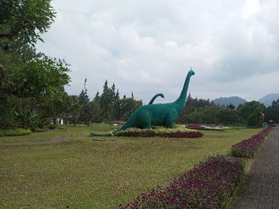 A large, green dinosaur-shaped shrub stands prominently in a park-like setting. The lawn is well-manicured, with borders of purple flowers lining a pathway. In the background, mountains are visible under a cloudy sky, with various trees and foliage adding to the scenic environment. A smaller, similar green dinosaur shrub is situated further into the background.