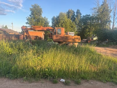 An orange excavator sits parked in a field of tall green grass under a clear blue sky. The excavator appears old and rusted. Behind the excavator, a weathered wooden fence encloses a small, dilapidated building. Further in the background, a line of trees stands tall, their green foliage contrasting with the sky. A smaller, orange tractor is visible in the distance, also parked. The scene is bathed in the warm light of late afternoon or early evening, casting long shadows and highlighting the texture of the grass. The overall atmosphere is one of stillness and abandonment, with nature beginning to reclaim the machinery and structures. There is no visible text in the image.