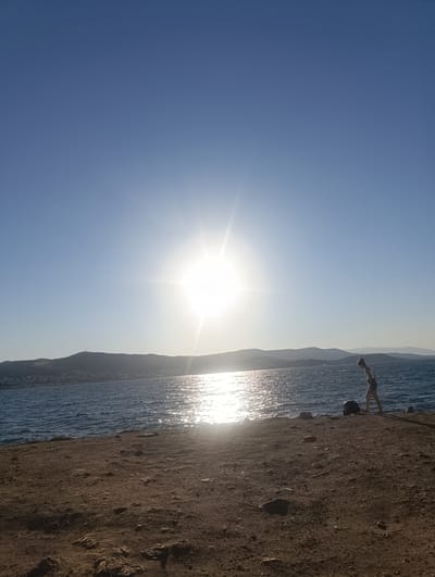 The image depicts a serene coastal scene bathed in the bright, warm light of the sun. A lone individual, a young person dressed in a white top and shorts, is walking near the water's edge. They appear to be bending down, perhaps interacting with something on the ground or the shoreline. Beside them, a dark, circular object, possibly a bag or a large rock, rests on the ground. The setting is a rough, sandy embankment leading down to a body of water with visible waves. Across the water, a silhouette of hills and a town is visible under the hazy glow of the sun. The sun itself is a prominent feature, casting a brilliant glare on the water and dominating the sky, suggesting it is either sunrise or sunset. The sky is a clear, pale blue, gradually transitioning to a brighter white near the sun. The overall mood is peaceful and contemplative, with the natural elements taking center stage. The location is identified as Urla, Turkey. There is no visible text in the image.
