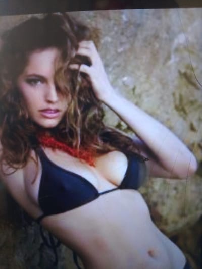 The image shows a medium shot of a woman with brown wavy hair, wearing a black bikini top and a red beaded necklace. Her hand is in her hair, and she is leaning against a rocky surface. The background is blurred and appears to be outdoors. The image quality is poor, with a low resolution and significant digital noise, making it difficult to discern fine details. There is no information available regarding the location in Dobra Krynytsia, Ukraine, or any other specific details about the scene or the subject.