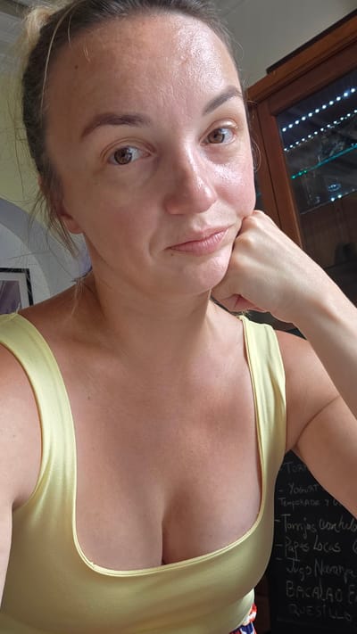 A medium shot captured from a slightly low angle shows a woman with blonde hair pulled back in a bun, her head tilted to her right. Her left hand rests under her chin, with her elbow bent and forearm resting against her jawline. She is looking directly at the camera with a neutral expression. She is wearing a light yellow top with wide straps, revealing her décolletage.
In the background, to the right, a dark wooden cabinet with glass doors is visible, illuminated by strings of white lights. The glass doors display shelves with various items, though they are out of focus. To the left of the frame, a portion of a framed picture or artwork can be seen.
A blackboard menu is partially visible on the right side of the image, displaying handwritten text in white. Some of the visible items include "TORTA," "YOGURT," "TEMPORADA Y Q," "Torrijas con Huevo," "Papas Locas," "Jugo Naranja," "BACALAO EN," and "QUESILLO."
The scene appears to be indoors, possibly a restaurant or cafe, given the menu and display cabinet. The overall lighting suggests it is daytime, with natural light possibly mixed with artificial illumination from the background cabinet. The image is underexposed in some areas, particularly the background.