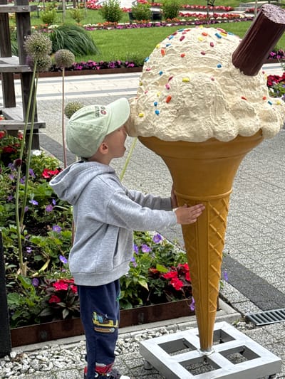 In Bruzdeilynas, Lithuania, a young boy wearing a gray hoodie and dark blue sweatpants adorned with cartoon construction vehicles is interacting with a large, oversized ice cream cone sculpture. The boy is leaning his head against the creamy white top of the ice cream, which is decorated with colorful sprinkles and a dark brown chocolate wafer. His hands are positioned on the golden waffle cone base of the sculpture. He wears a light green baseball cap. The sculpture is placed outdoors, with a backdrop of lush green grass, colorful flower beds featuring red and purple blooms, and decorative plant arrangements. The ground is a mosaic of gray paving stones and white gravel.