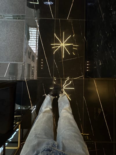 The image shows a person wearing blue jeans standing on a glossy black tiled floor with gold-colored veins or patterns. The floor reflects the surroundings, including a building with visible windows and a textured surface. The person's feet and shoes are visible, and part of a black chair is in the lower part of the frame. Based on metadata, the location is Ciudad López Mateos, Mexico.