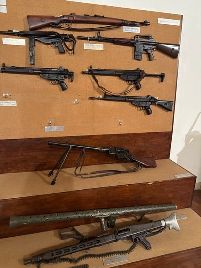 The image displays an indoor exhibit of military firearms within a museum setting in Praia do Ribatejo, Portugal. The scene features multiple rifles, submachine guns, and machine guns mounted on a two-tiered display system against an off-white wall.
The upper tier consists of a light brown corkboard backing, upon which eight firearms are horizontally secured with metal clips. From top to bottom, left to right:
* A bolt-action rifle with a wooden stock and a brown sling is positioned at the top.
* Below it, a German MP40 submachine gun with a folding stock and a black sling is visible.
* To the right of the MP40, an AR-pattern assault rifle with a dark stock, carrying handle, and a black sling is mounted.
* In the second row, two black-finished roller-delayed blowback rifles, consistent with H&K G3 models, are displayed; the left one with a solid stock, the right one with a solid stock and a black sling.
* In the third row, two more black-finished roller-delayed blowback rifles, also consistent with H&K G3 models, are displayed; the left one with a solid stock, the right one with a solid stock and a black sling.
* Small white rectangular labels with black text are present below several firearms, providing identification.
The lower tier consists of two dark wooden shelves.
* On the upper wooden shelf, a light automatic rifle with a wooden stock, an extended bipod, and a black sling, resembling a Chauchat, is displayed horizontally. A small white label is visible on the right side of this shelf.
* On the lower wooden shelf, a long, green, perforated tubular object, possibly a barrel shroud or a section of a recoilless rifle, is positioned above a black general-purpose machine gun, consistent with an MG 42 or MG 3. The machine gun features a bipod and a section of an ammunition belt is visible below its receiver. A small white label with black text is located below this machine gun.
No people are present in the image. The environment suggests a public display for historical or educational purposes.