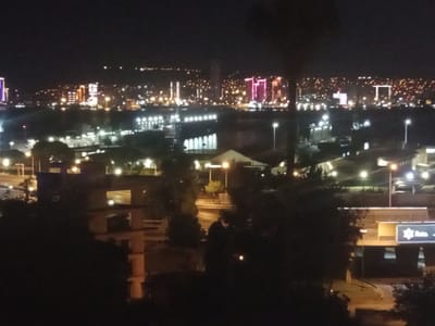 The image shows a nighttime view of the Bayraklı district in İzmir, Turkey. In the distance, a city skyline is illuminated by numerous lights from buildings. Some of these buildings are tall and appear to be skyscrapers, with distinct architectural features and colorful lighting, such as pink and blue lights on a prominent structure. Closer to the foreground, there are lower-rise buildings, some with illuminated windows and outdoor lighting. Streetlights are visible, casting pools of light on the ground. Darker silhouettes of trees and structures are present in the immediate foreground, partially obscuring the view. There are no discernible individuals or specific actions captured in the image, with the focus being on the urban landscape at night. The overall impression is of a bustling city environment at rest under the cover of darkness.
