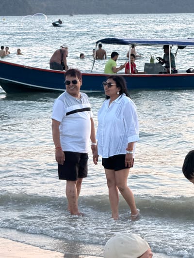 The image displays a daytime scene at Cenang Beach, Malaysia, featuring multiple individuals in and around the ocean.

In the foreground, a man and a woman stand side-by-side in shallow ocean water, approximately ankle to mid-calf deep, with small waves breaking around their bare feet. The man, on the left, wears a white short-sleeved t-shirt with a grey upper chest and a black horizontal stripe, dark shorts, and sunglasses. A silver watch and red string bracelets are visible on his left wrist. He smiles towards the right. His bare legs show several dark marks or spots. The woman, on the right, wears a white long-sleeved collared shirt over dark shorts, sunglasses, and white earrings. Her left wrist displays multiple white and red beaded bracelets along with a red string bracelet. She looks towards the left with a slight smile. The top of a beige cap, belonging to a partially cropped person, is visible in the bottom right corner.

In the midground, a blue boat with a white stripe along its hull and a blue canopy is positioned in the water behind the two primary subjects. Several individuals are on board, including one seated at the helm on the right, one in a red top towards the center, and another in a lime-green shirt on the left. Multiple other people are visible in the water, wading or swimming around the boat, including a man with a bare back to the left of the vessel.

The background shows open ocean with a distant landmass or hills on the horizon. A person on a jet ski is observed moving across the water towards the left. Two large, translucent bubbles float in the air above the water, to the left of the jet ski. The water surface is calm with minor ripples and small breaking waves near the shore.
