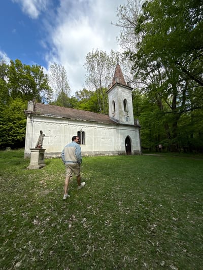 The image shows a man standing in front of the Chapel of the Snow Virgin Mary, located in the park of the Červený Kameň Castle in Častá, Slovakia. The chapel is a light-colored building with a red-tiled roof and a small bell tower. A statue of a religious figure stands on a pedestal in front of the chapel. The man is wearing shorts, a light-colored vest, and sunglasses. The environment is a green park with trees. The sky is visible with some clouds.