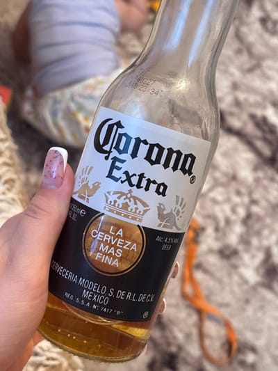 A person's hand with a white-tipped manicure holds a clear glass bottle of Corona Extra beer. The bottle's label is black with white text and features the brand name "Corona Extra," along with the phrase "LA CERVEZA MAS FINA" within a circular emblem. Below this, the label indicates "Alc. 4.5% Vol. BEER" and text identifying the manufacturer as "CERVECERIA MODELO, S. DE R.L. DE C.V. MEXICO REG. S.S.A. N° 7417 "B"." The beer inside the bottle is a golden amber color. The background is blurred, showing a person in striped clothing and a patterned blanket or rug. There is no discernible information about the location in Sovkhoza "Razdol'e", Russia.