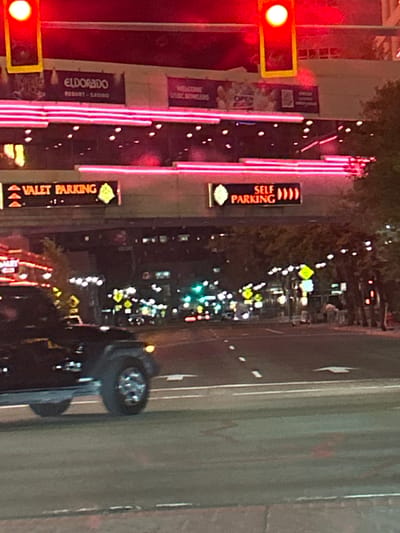 This image depicts a nighttime urban street scene in Reno, Nevada, facing the Eldorado Resort Casino. Two prominent red traffic lights with yellow housings are visible at the top. The background is dominated by the multi-story Eldorado Resort Casino building, identified by a large "ELDORADO RESORT - CASINO" sign and illuminated by extensive pink neon strip lighting. Additional signs on the building include "WELCOME USC BOWLERS" and "OPEN". Below the casino, an overhead structure supports two illuminated directional signs: "VALET PARKING" with three left-pointing arrows and "SELF PARKING" with multiple right-pointing symbols.

In the lower-left foreground, a dark-colored SUV, possibly a Jeep, is captured in motion, with its front passenger side and a chrome wheel visible, its headlight illuminated. The multi-lane asphalt road features white dashed lane markings and a white arrow marking further down on the right. Numerous streetlights illuminate the roadway and sidewalks extending into the blurred distance, where additional vehicles and green traffic signals are discernible. Trees with dark foliage line the right side of the street, and a few indistinct human figures are visible on the sidewalk in the far right mid-ground. The overall scene is heavily saturated with artificial light.