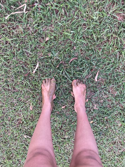 A first-person perspective photograph captures the lower legs and bare feet of an individual standing on a patch of grass. Both feet are visibly coated with a thick layer of dark brown, wet mud, extending over the toes and soles. The skin on the visible portions of the legs appears light brown. The ground consists of green grass interspersed with dry leaves and exposed areas of soil. The scene is located in Pirenópolis, Brazil.