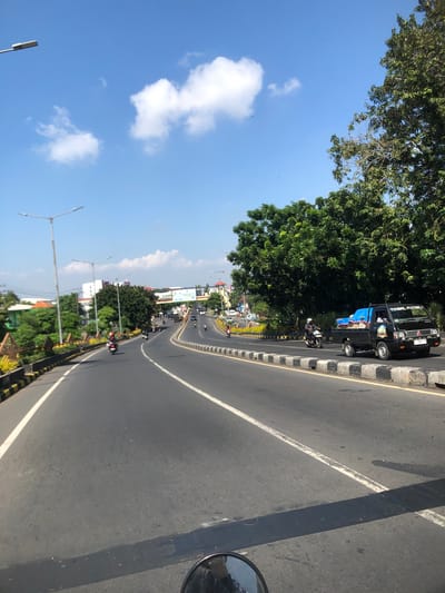 The image depicts a street scene in Buduran, Indonesia, under a clear blue sky with scattered clouds. The perspective is from a moving vehicle, likely a motorcycle, looking forward down a multi-lane paved road. On the right side of the road, a black pickup truck with a blue tarp covering its bed is visible, along with a motorcyclist beside it. Further down the road, more vehicles, including several motorcycles, are traveling in the same direction.

The road is flanked by a concrete median with black and white striped barriers. To the left of the road, there is a yellow and black striped barrier, followed by a landscaped area with plants and decorative elements. Beyond the landscaping, buildings and trees are visible. Tall streetlights with multiple bulbs are positioned along the road. In the distance, an overpass or bridge with signage is visible, spanning the road. The overall impression is of a moderately busy urban or suburban environment on a sunny day.