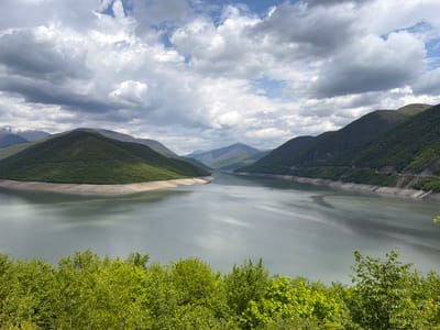The image depicts a landscape scene in the vicinity of Aranisi, Georgia, featuring a large reservoir bordered by mountains. The water surface is reflective, with subtle variations in color and texture, suggesting the presence of light wind. The mountains are covered in dense green vegetation, with some exhibiting evidence of a road system constructed along the steep slopes. Patches of snow can be seen on the peaks of more distant mountains. In the foreground, there is lush greenery. The sky is overcast with a mix of grey and white clouds.