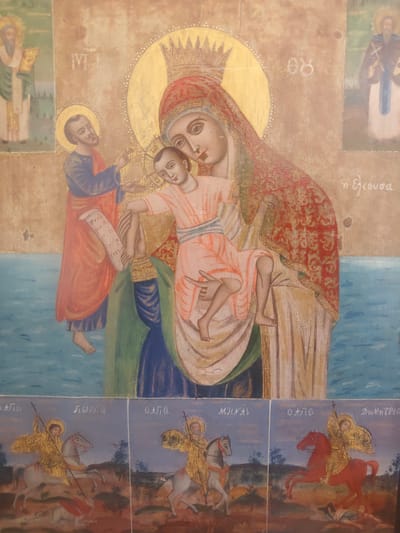 This is a religious icon depicting the Virgin Mary holding the Christ Child, with other figures present. The Virgin Mary is crowned and wears a red veil. The Christ Child is dressed in a pink tunic. To the left, a male figure in a blue and red robe holds a scroll. To the right, another robed figure is partially visible. In the background, there are depictions of saints or religious figures, one in a green robe and another in a brown robe with a blue sash. Below the main scene, there are three smaller panels, each showing a saint on horseback, wielding a spear, and battling a dragon or fallen figure. The style of the artwork suggests it is an Orthodox icon. The image is located in the Municipal Unit of Neapoli, Greece.
