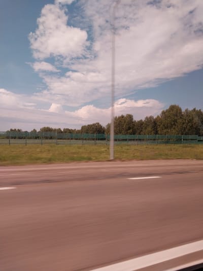 This is a photograph taken from a vehicle moving along a road in Nalobikha, Russia. The image is oriented vertically, with the right side of the road occupying the majority of the frame. The road surface is a light brown color and appears to be asphalt, with two white lane lines visible. The motion blur indicates the vehicle is in transit.
To the left of the road, a strip of green grass is visible, bordering a tall, green, chain-link fence. Beyond the fence, a dense line of trees forms a backdrop. The trees are a mix of leafy deciduous trees, appearing green and full, suggesting a summer or late spring setting.
Above the trees, a vast sky stretches out, filled with a mix of white, puffy clouds and streaks of lighter cirrus clouds against a bright blue background. The lighting suggests it is daytime, likely late morning or afternoon, with clear, fair weather. There is no visible text in the image. The scene evokes a sense of travel and the open road, with a peaceful, natural landscape to the side.