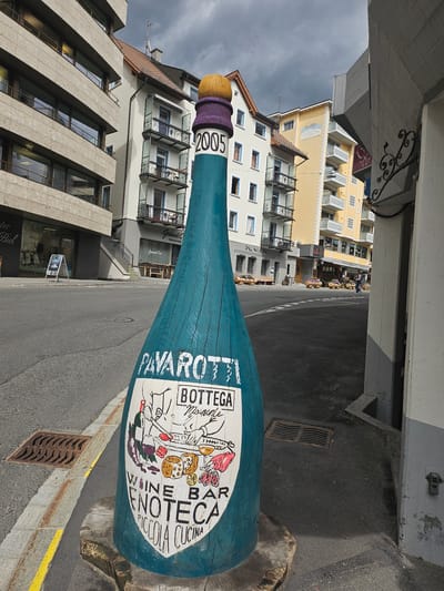 This is a photograph taken in St. Moritz, Switzerland, featuring a large, teal-colored wooden sculpture of a wine bottle, lying on its side on a street. The bottle is decorated with colorful illustrations and text. The text visible includes "2005", "PAVAROTTI", "BOTTEGA MAISONNE", "WINE BAR", "VENOTECA", and "PICCOLA CUCINA". The sculpture is situated on a street with a yellow painted line and a metal grate.
Behind the sculpture, several buildings are visible. The architecture suggests a European setting, with balconies and windows characteristic of alpine towns. One building is painted yellow and has the text "HEMMIG WAYS" and "Grand Cru" visible. Another building is lighter in color, with multiple floors and balconies. The overall scene appears to be a street in a town, possibly a commercial area given the signage on the buildings and the presence of an advertising sign in front of a store on the left.
The sky is overcast and appears to be a cloudy day. The lighting suggests it is daytime, but the weather might be cool or about to rain. There are no people prominently featured in the image, though there might be a few figures in the background of the yellow building. The focus is on the wine bottle sculpture and the surrounding urban environment.