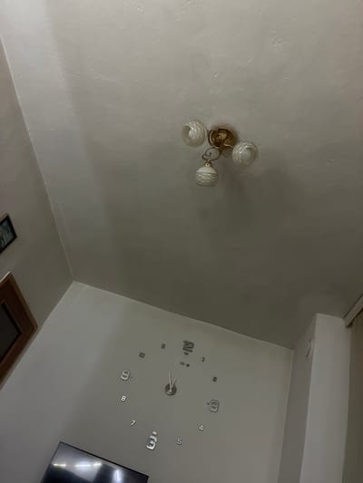 This is an indoor scene, reportedly located in the city of Vlahovo, Bulgaria, captured from a low, upward-angled perspective, showcasing a ceiling and portions of the upper walls.
The ceiling is uniformly light grey or off-white, characterized by a rough, uneven texture, indicative of plaster or stucco, with minor surface irregularities. A four-part ceiling light fixture is centrally mounted on the ceiling, featuring a gold-toned metallic base. Three of its arms support globe-shaped glass shades adorned with white, swirling decorative patterns; two shades extend upwards and outwards, while one hangs downwards. The light bulbs within each shade are visible.
The walls, also painted in a light grey or off-white, form visible corners within the room. On the upper left wall, a small, dark-framed picture with indistinct content is mounted high. Below it, a section of a wooden-framed object, possibly a window or a cabinet, is partially visible. The primary wall in the lower half of the image features a large, minimalist wall clock. This clock consists of individual, silver-colored, reflective Arabic numerals (1 through 12) adhered directly to the wall, with a central mechanism displaying hour and minute hands indicating approximately 11:59. On the far right, a vertical structural element, possibly a pillar or corner, is visible. At the very bottom center, a bright reflection suggests the presence of an electronic screen, likely a television, largely outside the frame. No people or active interactions are depicted.