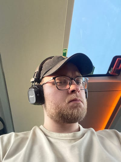 A person wearing glasses, a black baseball cap, and Marshall headphones is visible. The person has a light beard and is wearing a light-colored shirt. The scene appears to be inside a vehicle, possibly a train, as suggested by a window showing the sky and a partially visible emergency exit sign. Based on the location, this image could be from a train within Schipkau, Germany.