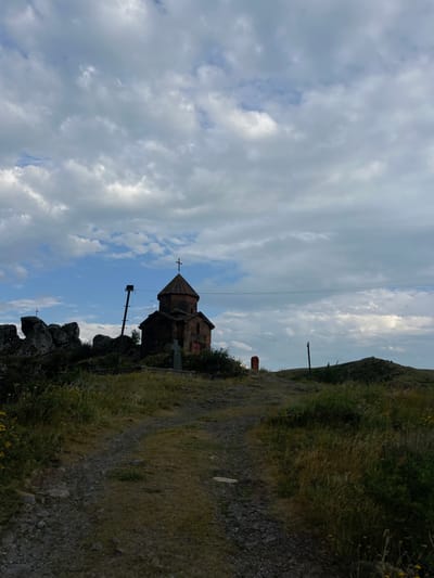 A stone church with a cross sits on a hillside, against a backdrop of cloudy skies. A dirt path winds alongside the church, lined with grass and rocks. Power lines stretch across the sky. The angle makes the scene appear skewed, with the sky on the left and hillside on the right. The architectural style of the church is traditional.