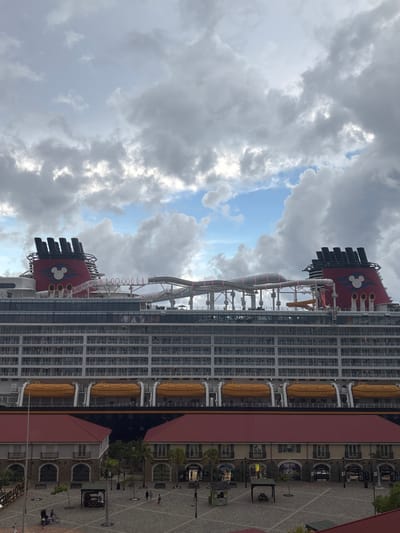 The image depicts a large Disney Cruise Line ship docked at the Falmouth cruise port in Falmouth, Jamaica. The massive vessel occupies the central and upper portions of the frame, towering over the foreground. It features multiple decks with visible cabins and balconies, and a row of orange lifeboats is mounted along its side. Two iconic red funnels with black tops and white Mickey Mouse silhouette logos are prominent on the upper deck. Between the funnels, a complex system of transparent water slides and tubes, including what appears to be the AquaDuck, is visible.

In the lower third of the image, the Falmouth cruise terminal plaza is visible, paved with a light-colored, patterned surface. Two buildings with red roofs, light-colored walls, and multiple arched doorways/windows flank the central plaza. Several smaller structures, including kiosks and covered seating areas, are dispersed across the plaza. Sparse vegetation, including palm trees, is present. Numerous individuals, identified as pedestrians, are seen walking and congregating within the plaza area. The sky above is partially cloudy, featuring a mix of grey, voluminous clouds and patches of clear blue sky. The perspective is elevated, looking down upon the dock area and up at the cruise ship.
