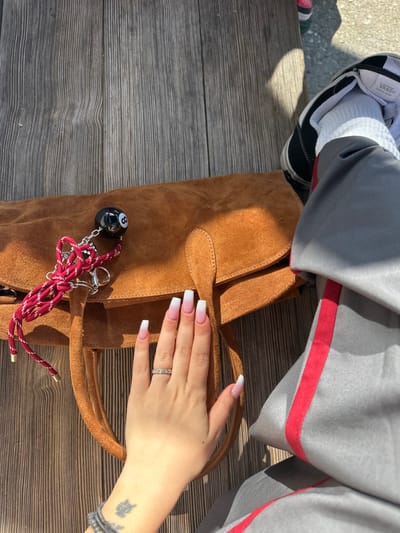 The image provides an overhead view of an outdoor scene in Glendale, United States, depicting personal items and portions of an individual.
A person's right hand, featuring French ombre acrylic nails, a silver-toned ring with five clear stones on the ring finger, a dark bracelet, and a dark tattoo on the inner wrist, rests near a brown suede handbag. Attached to the handbag's flap is a keychain assembly, comprising a black '8-ball' billiard charm and a red and white patterned cord with silver-toned metal hardware. To the right of the bag, a lower right leg and foot are visible, clad in gray trousers with a prominent red vertical stripe down the outer seam, white socks, and a black and white Vans Old Skool sneaker. All subjects and objects are situated on a light-toned wooden plank surface, exhibiting visible grain, consistent with an outdoor bench or table. The scene is illuminated by strong sunlight, casting distinct shadows.