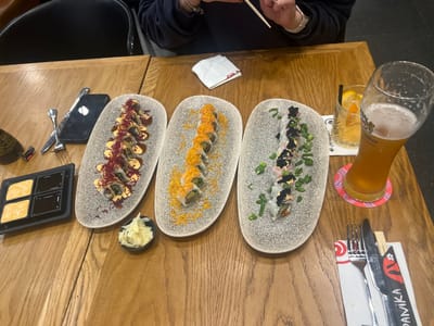 The scene depicts a dining table in a restaurant located in Kfar Yona, Israel. A light brown wooden table is set with various sushi dishes and beverages. In the upper portion of the image, the hands of one person are visible, holding white chopsticks over a sushi plate, wearing a dark long-sleeved garment and a silver-colored watch on the left wrist.
On the table, there are three elongated, speckled grey platters, each presenting ten pieces of sushi rolls. The leftmost platter features sushi rolls topped with a dark sauce, a lighter orange sauce, and red shredded garnish. The middle platter displays sushi rolls adorned with orange specks and light green garnish. The rightmost platter presents sushi rolls topped with black roe, yellow roe, and chopped green onions.
To the left, a black rectangular tray holds four compartments: two containing a light orange/yellow sauce and two with a dark brown sauce. Adjacent to this tray is a small dark brown bottle, likely soy sauce. A black smartphone is placed face down next to a metal knife and fork. A small black bowl of pickled ginger is positioned centrally.
On the right side of the table, a tall, fluted glass of light amber-colored beer with foam, bearing an "Elephant" brand logo, rests on a pink and purple coaster. Behind it, a shorter glass contains a light yellow drink with ice and an orange slice. Below these, a set of metal cutlery (knife and fork) is placed on a white paper napkin with red Japanese-style writing, "DAMIKA". A white folded paper napkin is also visible near the person's hands. The background indicates a dark floor and seating.