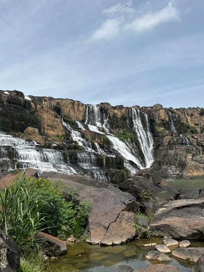 The image depicts the impressive Pongour Waterfall, located in Tân Phú, Vietnam. The waterfall cascades down a multi-tiered rock formation, creating a wide curtain of white water against a backdrop of rocky cliffs. The cliffs are a mix of earthy brown and darker, weathered rock, with patches of green vegetation clinging to their surfaces. At the base of the waterfall, several large, dark boulders are scattered around a pool of calm, greenish water. Lush green plants, including tall grasses and leafy bushes with small yellow flowers, grow in the foreground amongst the rocks, adding a vibrant touch to the scene. The sky above is partly cloudy with a pale blue hue.