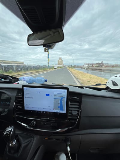 The image shows the interior of a vehicle, likely a van, looking forward through the windshield. The dashboard features a touchscreen infotainment system displaying navigation and radio information. The screen shows a map with the route highlighted, indicating a destination in Neuss, Germany. The radio is tuned to WDR Cosmo. A bottle of water is visible on the dashboard. Outside the vehicle, a road runs alongside a canal or river. In the distance, industrial buildings and cranes are visible under a cloudy sky. A white helmet is placed on the passenger side of the dashboard. The gear stick and part of the steering wheel are visible in the foreground.