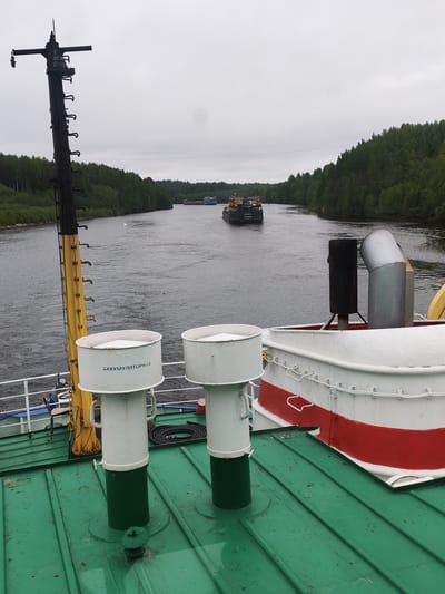 Captured from the deck of a vessel in Depo, Russia, the scene overlooks a waterway. Two barges are visible on the water, with tree-lined banks on either side. The overcast sky suggests a cloudy day. The foreground reveals details of the vessel's deck, including ventilation shafts marked with Cyrillic script ("АККУМУЛЯТОРНАЯ"), and other nautical equipment such as a mast and exhaust stacks. The deck is painted in green, white and red, with additional features of ropes and tubes visible.