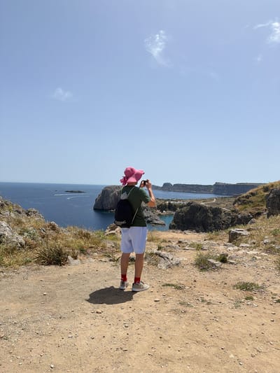 A person wearing a pink hat, dark green t-shirt, white shorts, red socks, and light-colored sneakers stands on a dirt path near a cliff's edge, taking a picture with a camera. The path is made of dry, light-brown dirt and scattered rocks. The person has a black bag slung over their shoulder.
Beyond the cliff's edge, there is a view of the blue sea with some rocky formations rising from the water. In the distance, the city of Lindos, Greece, can be seen along the coastline. The sky above is light blue with a few wispy clouds. The vegetation on the cliff edge is sparse and dry, with some small plants.