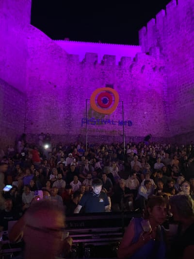 An outdoor nighttime concert is taking place against a stone wall illuminated with purple light. A circular logo with concentric orange and blue rings is displayed prominently on the wall, with the words "FESTIVAL MED" beneath it. A large audience is seated in rows facing a stage area, though the stage itself is not fully visible. Several people are standing near the front of the audience. A person in the foreground appears to be holding up a phone, possibly to take a picture or record the event. The overall atmosphere suggests a cultural festival or performance in a historic setting. The image is set in the city of Loulé, Portugal, likely within the castle walls which are characteristic of the city's historic center.
