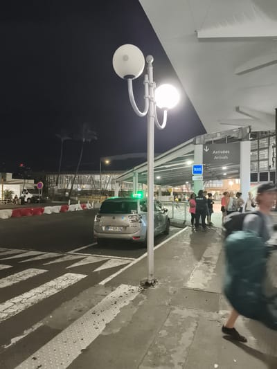 The image shows a nighttime scene at an airport terminal in Sainte-Marie, France, likely the Roland Garros Airport, given its location on Réunion Island, which is a French territory. A silver Citroën taxi, license plate FP-526-ZB, is parked on the right side of a pedestrian crossing. The taxi has a green illuminated sign on its roof, indicating it is operational.
Adjacent to the taxi is a sidewalk with a sign pointing downwards indicating "Arrivées Arrivals" and a smaller blue sign for "TAXIS". Several people are standing and waiting near the taxi stand, including a police officer wearing a uniform with "POLICE NATIONALE" written on the back. Others are dressed in casual attire.
In the background, palm trees are visible against a dark sky. The airport terminal building features modern architecture with a large, covered overhang illuminated by lights. The ground is wet, suggesting recent rain or cleaning. A lamppost with two globe-shaped lights stands prominently in the foreground. A person wearing a backpack is walking away from the camera on the right side of the frame, their movement blurred. The overall impression is one of an active but somewhat subdued transportation hub at night.