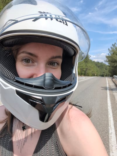 A woman wearing a white helmet with a clear visor is taking a selfie while riding a motorcycle. She has brown hair and green eyes and is wearing a grey top and a silver necklace. The road behind her is paved and lined with trees. The sky is blue with scattered clouds. The image appears to have been taken on a sunny day. The location is identified as Dzhankhot, Russia.