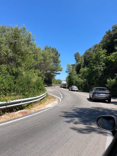 The image shows a winding road in a wooded area, likely in the vicinity of Mattinata, Italy. A silver sedan is in the foreground, driving on the road. Further ahead, another silver sedan is visible, as well as a white tanker truck. The road is bordered by lush green trees and bushes, with a metal guardrail on the left side of the road. The sky is a clear blue with a few faint white wisps. The overall scene suggests a scenic drive through a natural landscape.