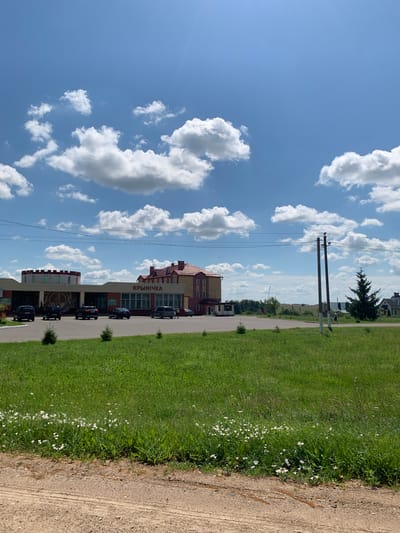 The image depicts a sunny day in Слабада-Кучынскі сельскі Савет, Belarus, with a bright blue sky dotted with fluffy white clouds. The foreground is a dirt road transitioning into a vibrant green grassy field, sparsely populated with small white flowers and saplings.
In the midground, a commercial building complex is visible. One section features a round, tower-like structure with a red trim. Adjacent to it is a longer, single-story building with a tiled red roof. Above the entrance of this section, the Cyrillic text "КРЫНІЧКА" is prominently displayed. Several cars are parked in front of these buildings, including a dark SUV, a black sedan, a dark hatchback, and another dark sedan.
Further to the right, a two-story building with a red-tiled roof and yellow siding stands, suggesting it may be part of the same complex or a neighboring establishment. A tall, dark green evergreen tree is positioned to the right of this building. Two utility poles with wires strung between them are also noticeable. In the far background, a flat landscape with trees and some rural structures can be seen. A person is walking towards the right of the frame near the utility poles, and another person is standing near the evergreen tree.