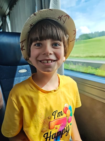 A young boy wearing a straw hat and a yellow t-shirt with "I'm 7 Happy" printed on it, sits in a bus seat. He is smiling at the camera. The background shows a window with a view of green fields and blue sky. The image was taken on the A1 West Autobahn in Eugendorf, Austria.