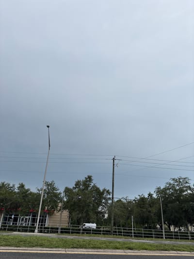 The image depicts a cloudy sky over a street scene in Bradenton, Florida, United States of America. In the foreground, a paved road with yellow and white lines is visible. Beyond the road, a grassy median separates it from a sidewalk and a white metal fence. Behind the fence, a row of trees lines the property. A light pole stands prominently in the middle left of the frame. Further back, a building with a tan facade and a red awning is partially obscured by the trees, with several windows visible. A white van is parked in front of the building. Another light pole and a utility pole with power lines are also present in the scene, spanning across the sky. The overall atmosphere is overcast and somewhat dull, indicative of a cloudy day.