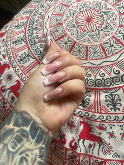 The image shows a person's hand with long, acrylic French manicured nails resting on a fabric with a red, black, and white folk art pattern. The pattern features circular mandalas and stylized horses and birds. The person's arm, visible in the lower left of the image, has a dark, intricate tattoo. The fabric appears to be a tablecloth or a similar textile. The location is not specified as being in Коробицыно, Russia; this is an assumption.