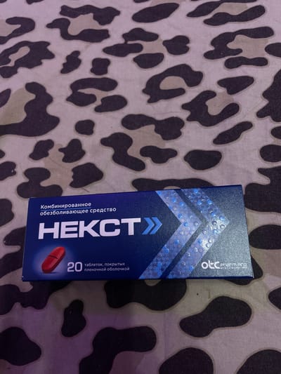 The image shows a close-up, overhead view of a blue box of medication placed on a patterned fabric. The fabric has a camouflage-like pattern with dark and light brown shapes against a lighter background. The medication box is dark blue with white text that reads "Комбинированное обезболивающее средство" (Combined analgesic agent) and "НЕКСТ" (NEXT) in large, bold white letters. Below "НЕКСТ", there is an image of a single red pill, followed by the number "20" and the text "таблеток, покрытых пленочной оболочкой" (tablets, film-coated). In the lower right corner of the box, a logo reads "otc Pharm PRO". There is no visible indication of the location being Vaganovo, Russia, or any people or actions taking place.