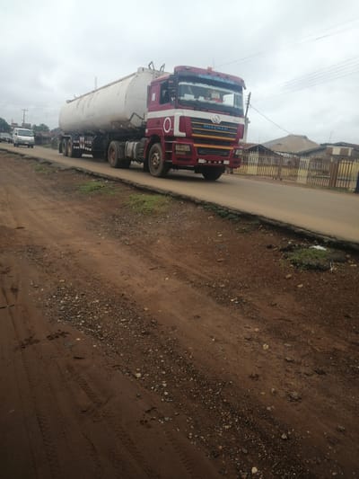 A wide shot captures a white tanker truck with red accents driving on an asphalt road in Ajasse Ipo, Nigeria. The truck's cab is visible, with "SHACMAN" printed on the front. The tanker is long and cylindrical. To the left of the truck, a white van is parked on the side of the road. The environment includes a dirt shoulder with tire tracks and footprints, scattered rocks, and some sparse green vegetation. In the background, there are fences, buildings with corrugated roofs, and utility poles with overhead wires against a cloudy sky. The overall scene depicts a typical roadside environment in a Nigerian town.