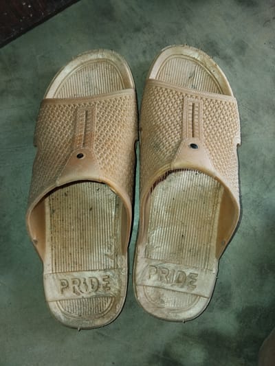 The image shows a pair of light brown, worn-out flip-flops placed on a greyish-green concrete floor. The flip-flops are a size large and appear to be made of rubber or a similar material. They have a textured pattern on the upper straps and a ribbed sole. The word "PRIDE" is embossed on the heel of each flip-flop. The floor is mottled and appears to be somewhat dirty, with some small dark specks visible on the flip-flops and the floor. In the top left corner, a dark wooden surface is visible. There are no people or any other objects in the immediate vicinity. The lighting suggests an indoor or shaded outdoor environment. The image provides no information about the location in Chhina Uttar, Pakistan.