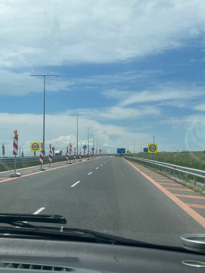 The image depicts a highway in Belokopitovo, Bulgaria. The view is from inside a moving vehicle, looking down the road. The road is divided by solid white lines with striped orange and white markers on either side. There are several signs indicating a speed limit of 100, as well as other road signs and streetlights. The sky is blue with scattered white clouds. There is a barrier on the side of the road. The vehicle's dashboard and windshield wipers are visible in the foreground.