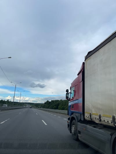 A truck is positioned on a highway near Бавыкино, Russia. The truck is red and blue. The sky is cloudy. The highway has multiple lanes and white dashed lines. Streetlights line the side of the road. Trees are visible in the background.