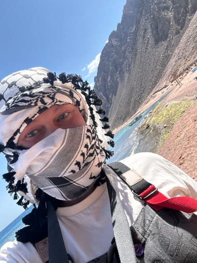 A person wearing a white and black keffiyeh covering most of their face is photographed in what appears to be a selfie. The person has light blue eyes. They are wearing a white shirt and a grey jacket with a purple button. A red and black strap is visible over their clothing. The background shows a landscape with mountains, a body of water, and a blue sky. Based on the location, it can be identified as being taken in Dahab, Egypt. The specific landmark is unidentifiable.
