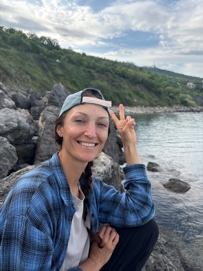 A woman with fair skin, wearing a blue plaid shirt, black pants, and a blue baseball cap, poses for a photograph outdoors. She is seated on a rocky outcrop overlooking a body of water, possibly the sea. In the background, a tree-covered hillside slopes down to the water's edge, and the sky is partially cloudy. The woman is smiling and holding up two fingers in a peace sign. The specific location is Bondarenkovo, Ukraine.